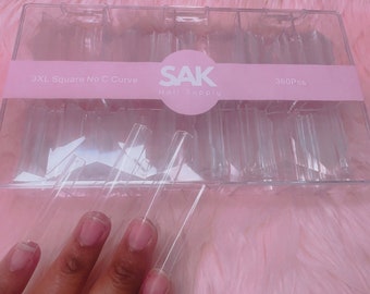 Könnte beinhalten: Eine transparente Plastikbox gefüllt mit 360 transparenten Plastik-Nagelspitzen. Die Box hat ein rosa Etikett mit der Aufschrift "3XL Square No C Curve SAK Nail Supply 360Pcs".