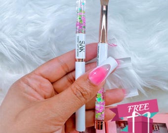 Könnte beinhalten: Zwei Nagelkunstpinsel, einer mit einem durchsichtigen Griff, der mit buntem Glitzer gefüllt ist, und der andere mit einem weißen Griff und einem braunen Pinselkopf. Die Pinsel werden in einer Hand mit rosa und weißen Ombre-Nägeln gehalten. Der Text "SAK SWEET ART KANA" ist auf dem Griff des Glitzerpinsels aufgedruckt. Der Text "FREE GIFT" ist in einer roten Box mit einem weißen Band aufgedruckt.