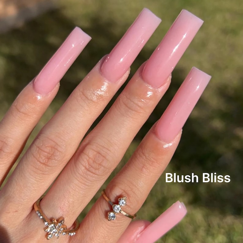 Puede incluir: Primer plano de una mano con u&ntilde;as acr&iacute;licas largas, cuadradas y de color rosa. Las u&ntilde;as tienen todas la misma longitud y un acabado liso y brillante. El texto "Blush Bliss" es visible en la esquina inferior derecha de la imagen.