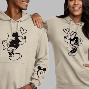 Könnte beinhalten: Zwei beigefarbene Hoodies mit schwarzen Mickey Mouse und Minnie Mouse Grafiken. Der Hoodie links zeigt Mickey Mouse, der Minnie Mouse küsst, mit einem Herzen darüber. Der Hoodie rechts zeigt Minnie Mouse, die Mickey Mouse küsst, mit einem Herzen darüber. Der Hoodie links hat ein Mickey Mouse Kopf-Motiv auf dem linken Ärmel.