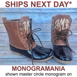 Può includere: Stivali da anatra marroni e neri con monogramma. Gli stivali hanno lacci marroni, occhielli e una tomaia in pelle marrone. La parte inferiore dello stivale è nera e lucida. I testi "SHIPS NEXT DAY*" e "MONOGRAMMANIA" sono visibili.