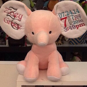 Op de afbeelding: Roze pluche olifant met witte oren. De oren zijn geborduurd met "Zoey Grace" op het ene oor en "07.24.14 7 pond 7 oz 20 inch 3:49pm" op het andere oor.