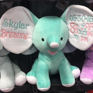 Op de afbeelding: Een lichtblauwe pluche olifant met witte oren. De oren zijn geborduurd met de tekst "Skylar Breanne" en "08:30:16 8 lbs 4 oz 20.75 in 7:19 PM".
