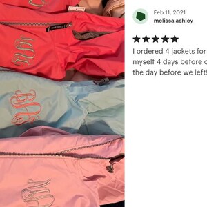 May include: Three rain jackets in bright coral, light blue, and soft pink, each featuring a silver-toned zipper and a decorative embroidered monogram on the chest. The jackets are layered vertically, showcasing the smooth fabric and stitching.