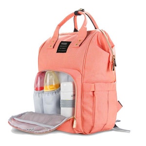 K&ouml;nnte beinhalten: Ein pfirsichfarbener Wickeltasche-Rucksack mit goldenen Akzenten und einem durchsichtigen Fach auf der Vorderseite. Die Tasche hat mehrere Taschen und F&auml;cher, darunter ein Rei&szlig;verschlussfach auf der Vorderseite und ein gro&szlig;es Hauptfach. Die Tasche ist au&szlig;erdem mit verstellbaren Tr&auml;gern und einem Griff ausgestattet.