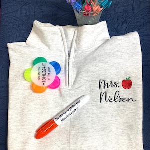 May include: A grey sweatshirt with a zip and a red apple embroidered on the front. The text "Mrs. Welsen" is embroidered below the apple. A rainbow flower button with the text "You are the Highlight of the year" is pinned to the sweatshirt. A white dry-erase marker with the text "The best way to predict your future is to create it" is lying on the sweatshirt.