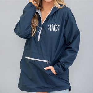 May include: Navy blue anorak jacket with a hood and a half-zip closure. The jacket features a white monogram on the front and a front pocket with a zipper. The interior lining is grey. The jacket has a drawstring at the bottom.