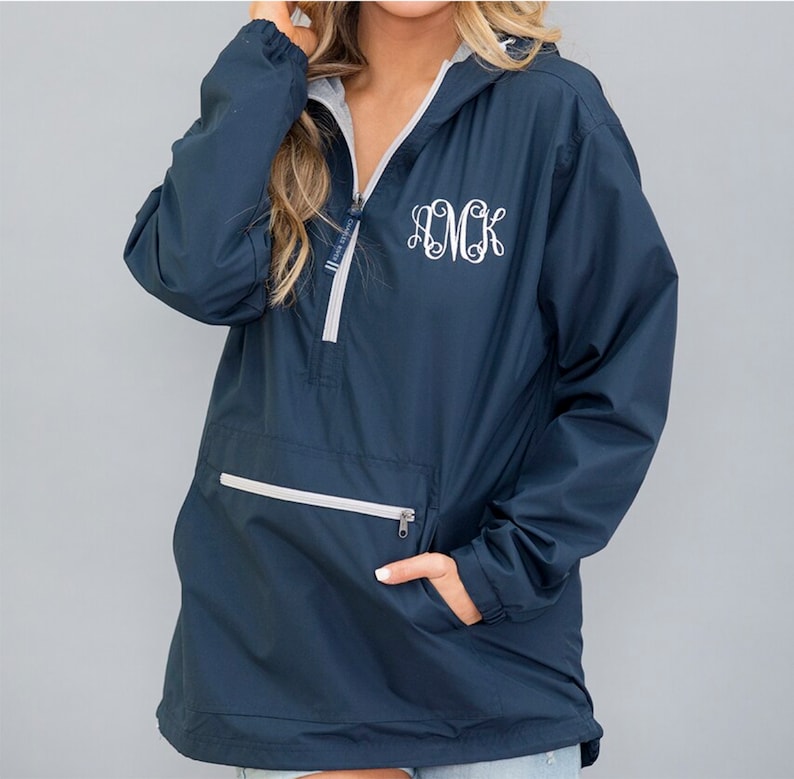 May include: Navy blue anorak jacket with a hood and a half-zip closure. The jacket features a white monogram on the front and a front pocket with a zipper. The interior lining is grey. The jacket has a drawstring at the bottom.