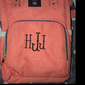 May include: A coral colored diaper bag with a zippered front pocket and a black embroidered monogram "HJL".