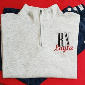 Könnte beinhalten: Ein grauer Quarter-Zip-Pullover mit einem schwarzen gestickten Monogramm "RN" und dem Namen "Layla" in roter Stickerei.
