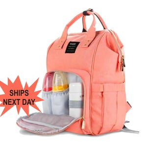 K&ouml;nnte beinhalten: Ein korallenfarbener Wickeltasche-Rucksack mit mehreren Taschen und einem transparenten Fenster auf der Vorderseite. Die Tasche hat ein Rei&szlig;verschluss-Hauptfach, eine gro&szlig;e Fronttasche und zwei Seitentaschen. Die Tasche ist au&szlig;erdem mit einer gepolsterten R&uuml;ckenpartie und verstellbaren Schultergurten ausgestattet.