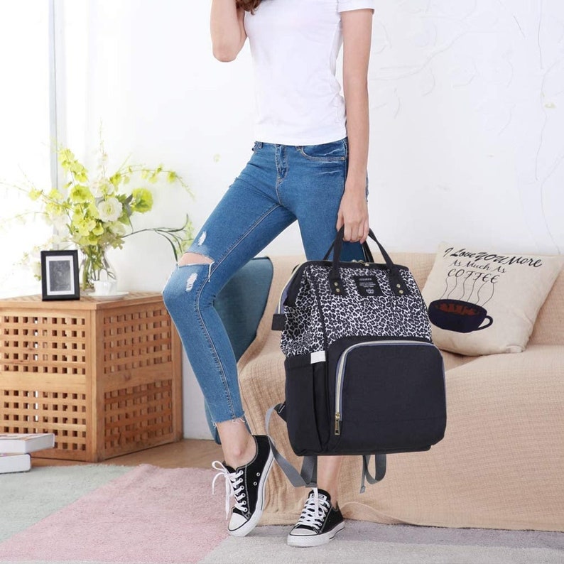 May include: A black and white leopard print diaper bag with a black bottom and gold accents. The bag has multiple pockets and a zipper closure. The bag is being held by a person wearing jeans and a white shirt.