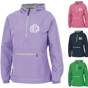May include: A lilac anorak jacket featuring a white monogram. The jacket has a hood, a front pocket with a zip, and a half-zip closure. Additional jackets in pink, navy blue, and green are also shown.