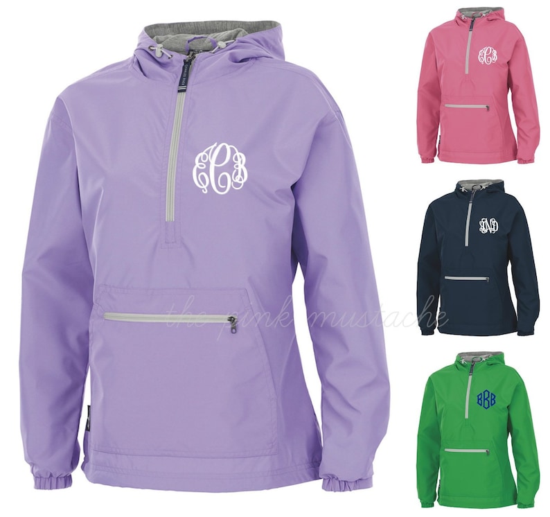 May include: A lilac anorak jacket featuring a white monogram. The jacket has a hood, a front pocket with a zip, and a half-zip closure. Additional jackets in pink, navy blue, and green are also shown.