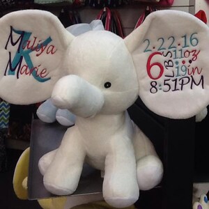 Op de afbeelding: Witte pluche olifant met blauwe oren. E&eacute;n oor is geborduurd met de naam "Maliya Marie" en het andere oor is geborduurd met de datum "2-22-16", "6 lbs 11 oz", "19 in" en "8:51 PM".