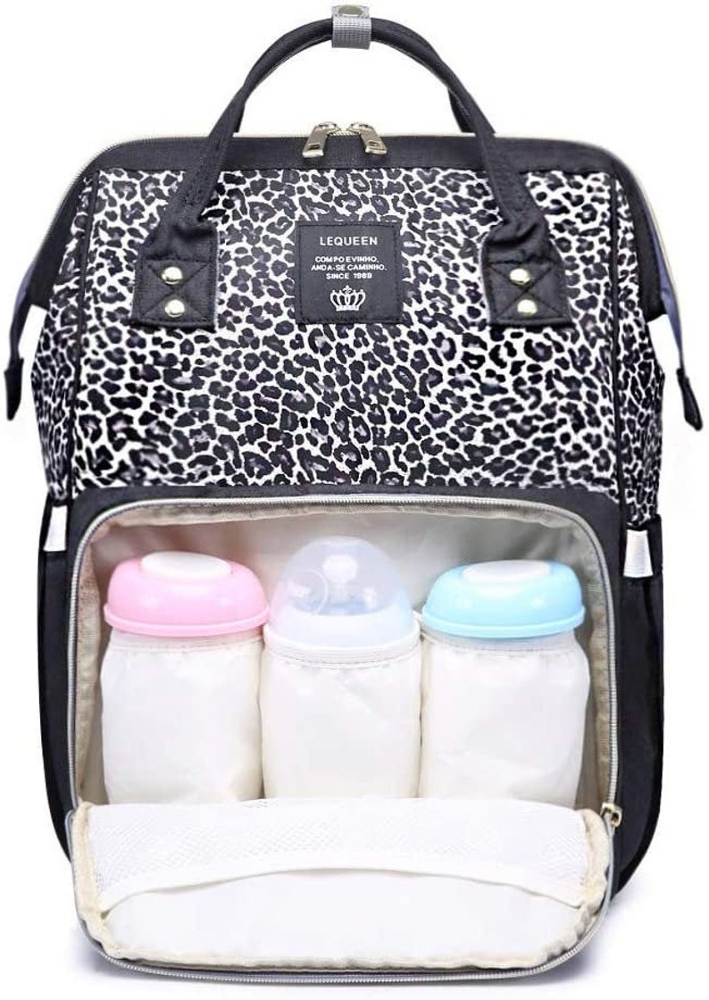 May include: A black and white leopard print diaper bag with a zippered top and multiple pockets. The bag has three insulated bottle holders and a mesh pocket inside. The bag has a black handle and a black adjustable shoulder strap. The bag has a label that says "LEQUEEN COMPO EVINHO, AMADA-SE CAMINHO, SINCE 1989".