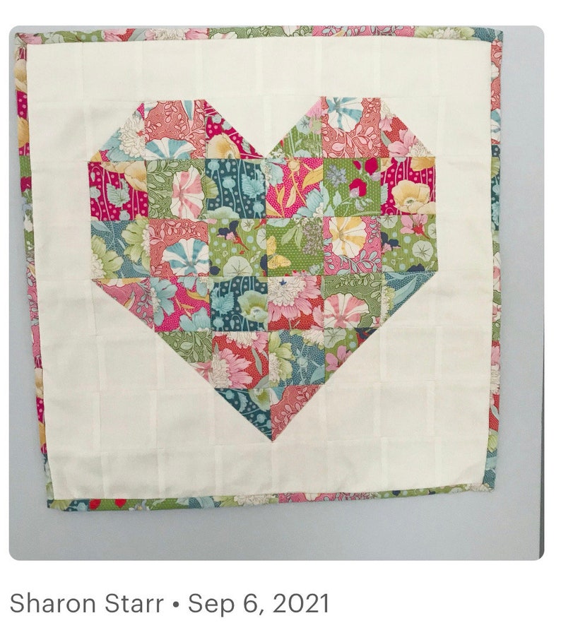 May include: A white fabric square with a patchwork heart design in shades of pink, green, and blue. The heart is made of small squares with floral patterns. The fabric is quilted and has a white border.
