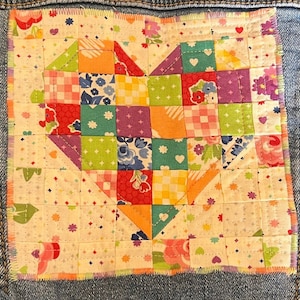 May include: A patchwork heart made of colorful fabric squares is sewn onto a denim jacket. The heart is made of various floral and geometric patterns in shades of pink, red, orange, yellow, green, blue, and purple.