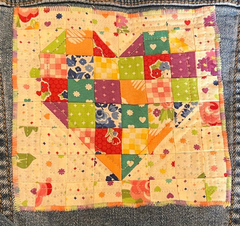 May include: A patchwork heart made of colorful fabric squares is sewn onto a denim jacket. The heart is made of various floral and geometric patterns in shades of pink, red, orange, yellow, green, blue, and purple.