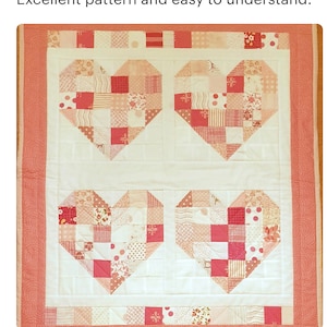 May include: A quilt with a white background and four pink hearts made of patchwork fabric. The hearts are made of various shades of pink, red, and white fabrics with different patterns, including polka dots, stripes, and floral prints. The quilt is bordered with a solid pink fabric.