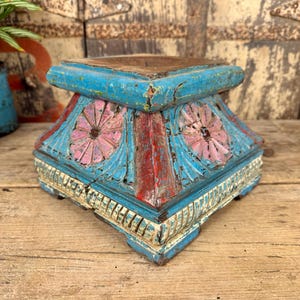 May include: A distressed, square wooden pedestal painted in shades of blue, red, and pink. The pedestal features carved floral designs and decorative trim. The aged appearance suggests it is an antique or vintage decorative item.