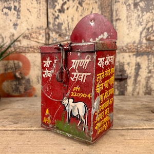 May include: A distressed red metal box with a hinged lid and latch. The box features painted text and a graphic of a white cow. The box has a rectangular shape and a small, curved top. The box shows signs of wear and tear.