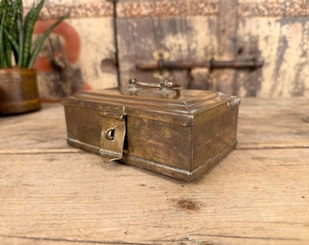 Puede incluir: Una pequeña caja rectangular de latón con tapa con bisagras y cierre de cierre. La caja tiene un aspecto deslustrado y envejecido, lo que sugiere que es vintage. El fondo incluye una superficie de madera y un vistazo a una planta en maceta.