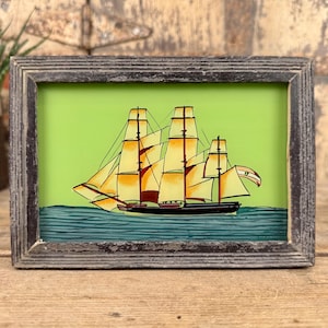 May include: A framed picture of a sailing ship with yellow sails and a black hull, set against a light green background. The artwork is framed in a weathered grey wooden frame, giving a vintage feel.