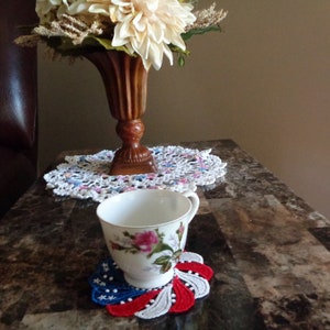 May include: A white porcelain teacup featuring a floral rose pattern, resting on a circular crocheted coaster with red, white, and blue star-shaped accents. Behind the cup sits a brown fluted vase holding cream-colored flowers on a white doily.