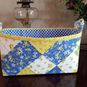 May include: A blue and yellow quilted storage basket with lemon and flower patterns. The basket has two handles and a gingham lining.