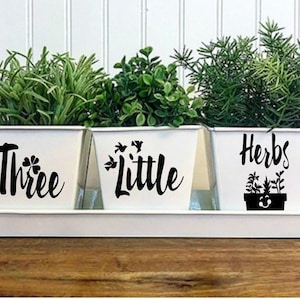May include: Three white metal planters with black lettering. The planters are labeled "Three", "Little", and "Herbs". The planter labeled "Herbs" has a small black and white illustration of a potted plant.