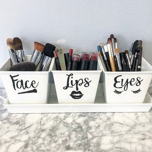May include: Three white metal containers with black labels and icons. The containers are labeled "Face", "Lips", and "Eyes". The containers are filled with makeup brushes, lipsticks, and eyeliner.
