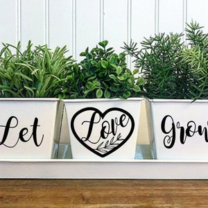 May include: Three white metal planters with black lettering. The planters are labeled "Let", "Love", and "Grow". The planters are sitting on a white metal tray.