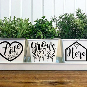 May include: A white metal planter set with three rectangular pots on a matching tray. Each pot has a black graphic and text: "Love" inside a heart, "Grows" with flower stems, and "Here" inside a house shape. The pots contain green plants.