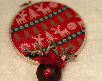 May include: A brown Christmas bauble with a reindeer face, red nose, and glitter eyelashes. The bauble is decorated with a red flower crown and brown antlers.