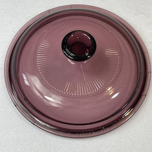 Puede incluir: Una tapa redonda y transparente de Pyrex de color púrpura con un pomo de color púrpura oscuro. La tapa tiene un diseño acanalado que irradia desde el centro. El borde y el pomo son de un tono púrpura más oscuro.
