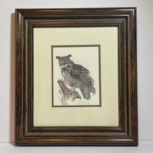 May include: A framed watercolor painting of a great horned owl perched on a tree branch. The owl features brown, gray, and white feathers with yellow eyes. The artwork is set within a cream-colored mat and a dark brown, textured wooden frame.