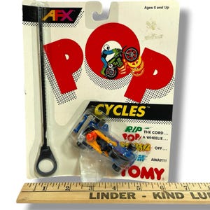 Puede incluir: Un juguete vintage AFX Pop Cycles, empaquetado en una tarjeta blanca y negra. La tarjeta presenta un diseño "POP" rojo con un gráfico de motocicleta. La motocicleta de juguete es azul y amarilla. El paquete incluye un cordón. A partir de 6 años.