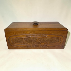 Puede incluir: Una panera rectangular de madera con acabado marrón oscuro y una tapa con un pomo central. El panel frontal muestra una escena en relieve de figuras en un campo con el texto Give Us This Day Our Daily Bread tallado en la madera.