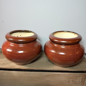 May include: Two matching ceramic bowls with a rounded, bulbous shape and a glossy, speckled reddish-brown glaze. Each bowl features a thick, smooth rim and a cream-colored interior, with small drainage holes visible along the inner wall.