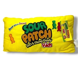 May include: A rectangular yellow plush pillow featuring the Sour Patch Kids logo in green and orange text. The pillow includes the phrase Sour then Sweet, small candy-shaped silhouettes in orange, green, and red, and a green product tag attached.