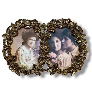 May include: A double picture frame made of gold-toned metal with an ornate, raised floral and leaf border. The frame holds two separate oval-shaped vintage-style photographs, one depicting a person with flowers and the other showing two people.