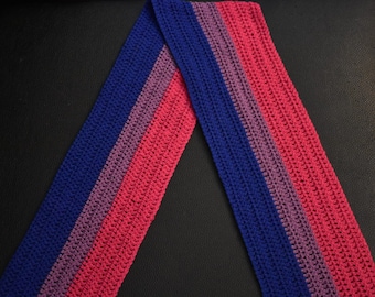 May include: A crocheted scarf with blue, purple, and pink stripes. The scarf is folded in half and laid flat on a black surface.