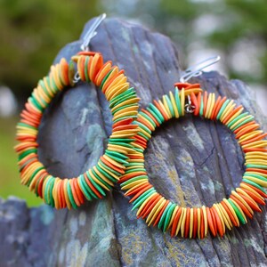 Könnte beinhalten: Ein Paar Creolen aus bunten, runden, flachen Perlen. Die Perlen sind in einem kreisförmigen Muster angeordnet und sind grün, orange und gelb. Die Ohrringe hängen an silbernen Haken.