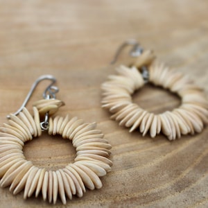 May include: A pair of earrings made of small, light brown, oval-shaped pieces of wood arranged in a circle. The earrings are attached to silver hooks.