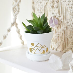 May include: White ceramic planter with a gold mountain and tree design. The planter is filled with a green succulent plant.