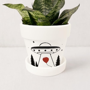 May include: White ceramic plant pot with a black illustration of a UFO, trees, and a red balloon. The pot contains a snake plant with green and white striped leaves. The pot sits on a matching saucer.