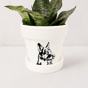 May include: White ceramic planter with a black silhouette of a German Shepherd dog. The planter has a matching saucer and is filled with a green snake plant.