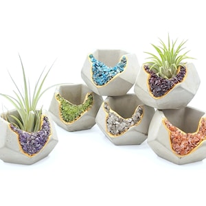 May include: Six geometric concrete planters with gold accents and various colored gemstone fillings. Each planter features a different gemstone, including amethyst, turquoise, green aventurine, clear quartz, and carnelian. Two planters have air plants growing inside.