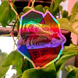 May include: A holographic sticker featuring a colorful snake skull with sharp teeth and the Japanese word "生ぜ" (pronounced "seze") meaning "to be born".